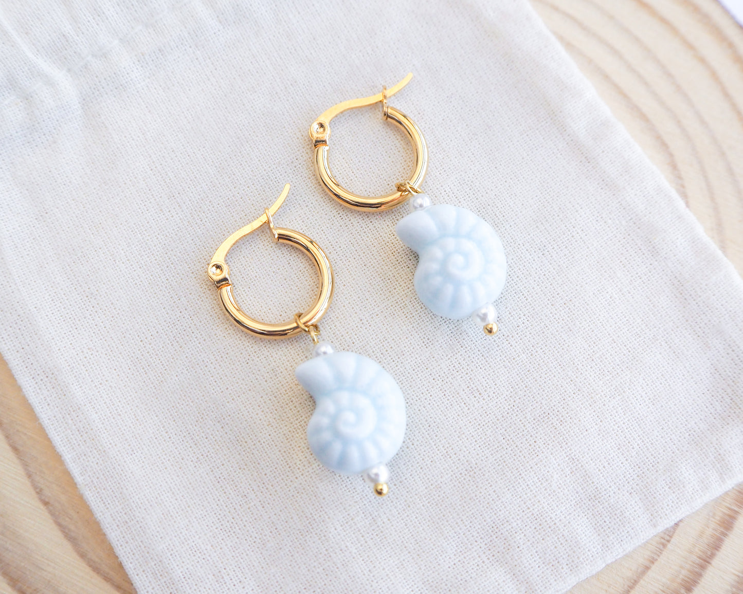 Gold ceramic seashell hoop earrings with light blue pherical charms on a white cloth background