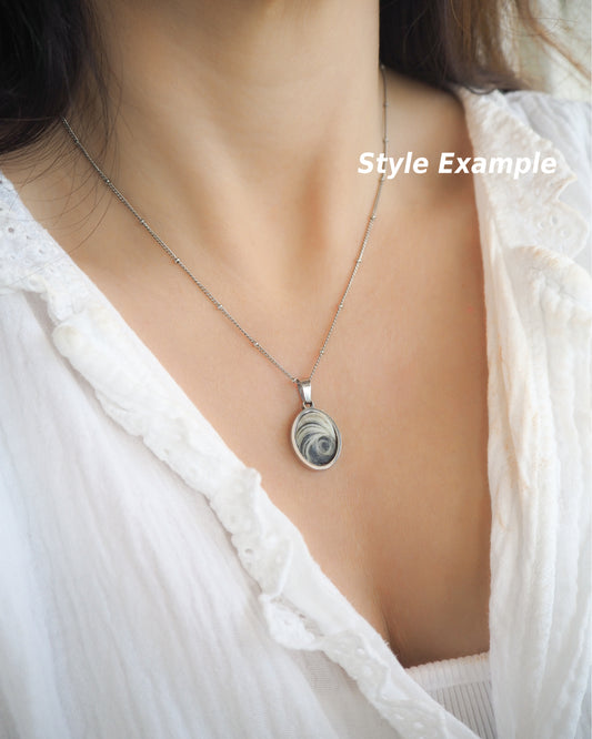Model wearing Dark Grey Eye of Saint Lucia Silver Necklace on neck with white blouse and brown hair, silver stainless steel shiva eye pendant necklace, Saint Lucy Eye, operculum pendant