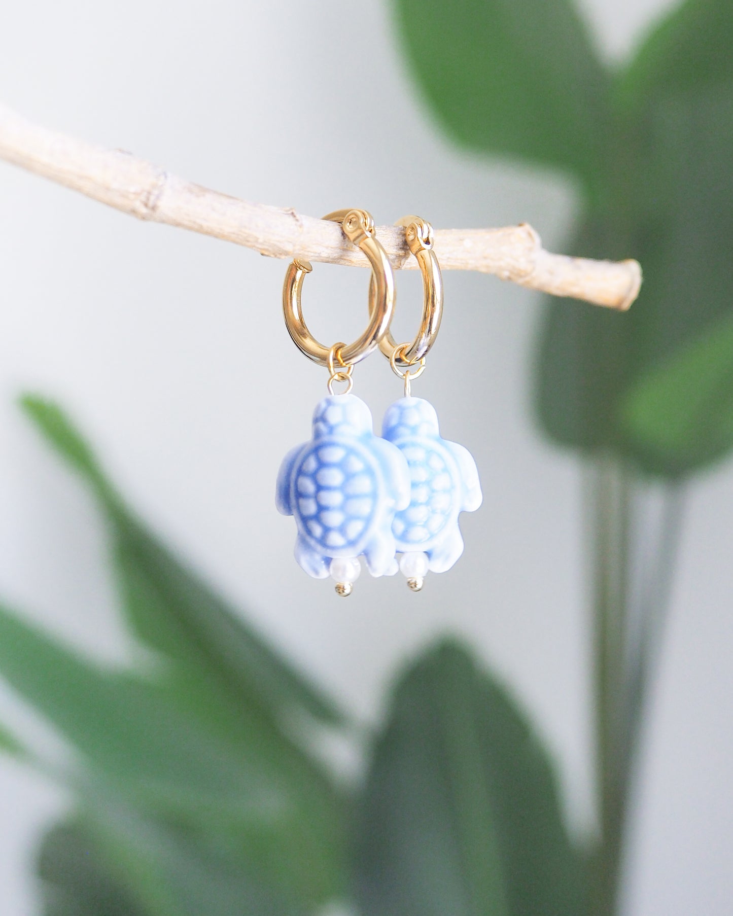 Gold hoop earrings with blue turtle charms on a branch with green leaves in the background