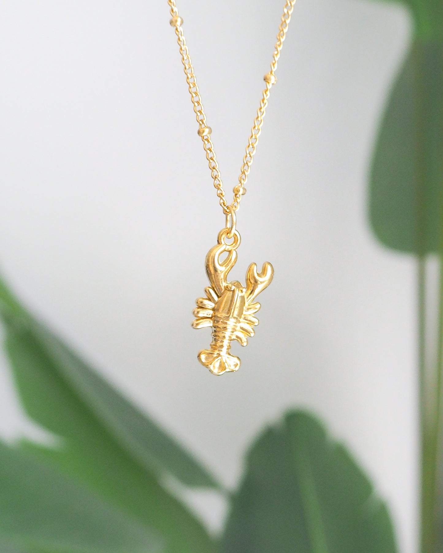 Front view of Gold Lobster Necklace with plant background, made with 18k gold stainless steel, coastal jewelry from Portugal