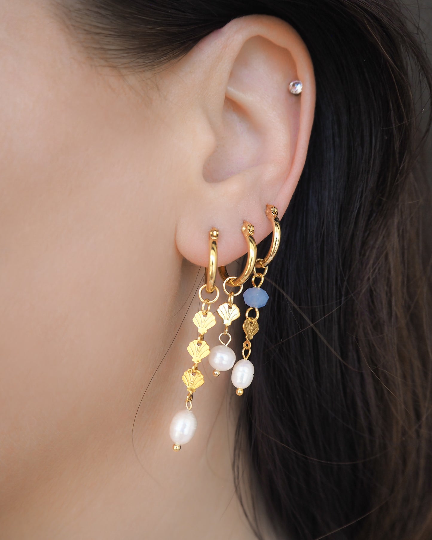 Model wearing mix of gold hoop earrings with blue and white beads and gold shell charms in ear. Handmade with blue sparkling crystal beads, ivory freshwater pearls and 14k gold stainless steel shell charms. The hoop earrings are made of 14k gold stainless steel These earrings are inspired on the ocean is part of the ocean serenity pearl collection.