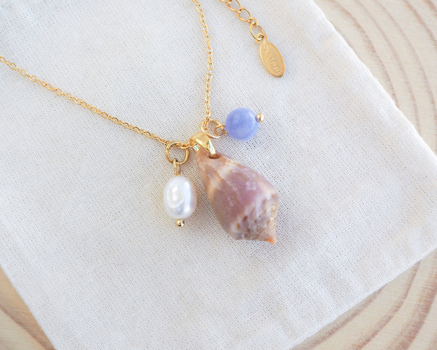 Close up of gold necklace with medium cone shell and a gemstone and pearl necklace on display. Mediterranean cone shell was handpicked from Portugal. Made with a ivory white freshwater pearl and blue angelite gemstone bead. Made with 18k gold stainless steel clasp and necklace chain.