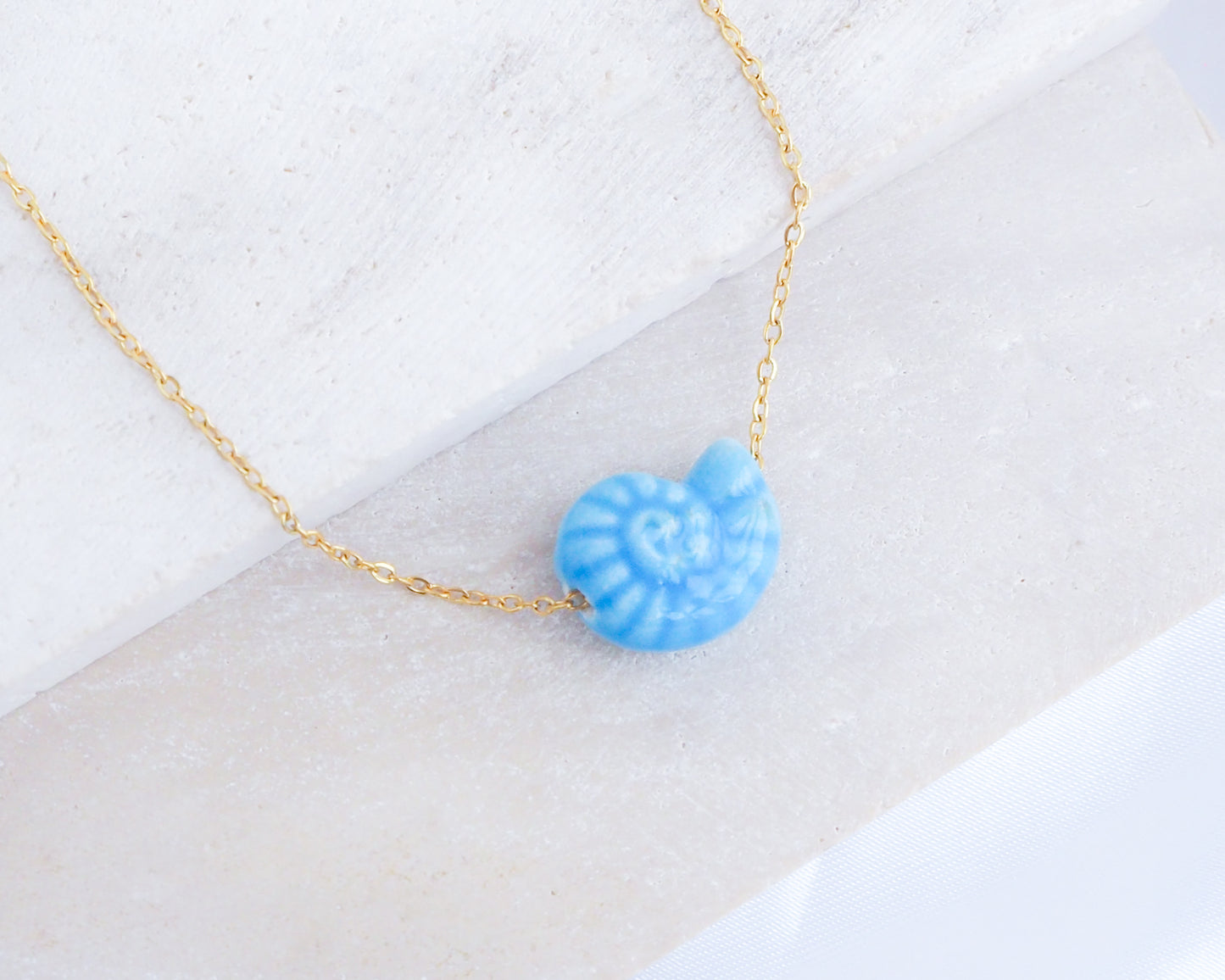 Mid Blue Ceramic Seashell Nautilus Gold Necklace displayed on stone