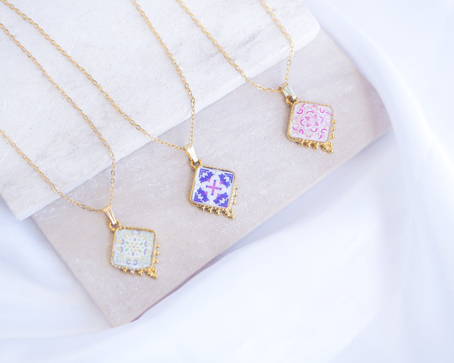 Mix of Portuguese Tile Necklaces on stone displayed. Made with 18k gold stainless steel and silver stainless steel. Showcasing white pink floral pendant, a green floral pendant and a purple pink floral pendant inspired by the Portuguese Azulejo design pattern.