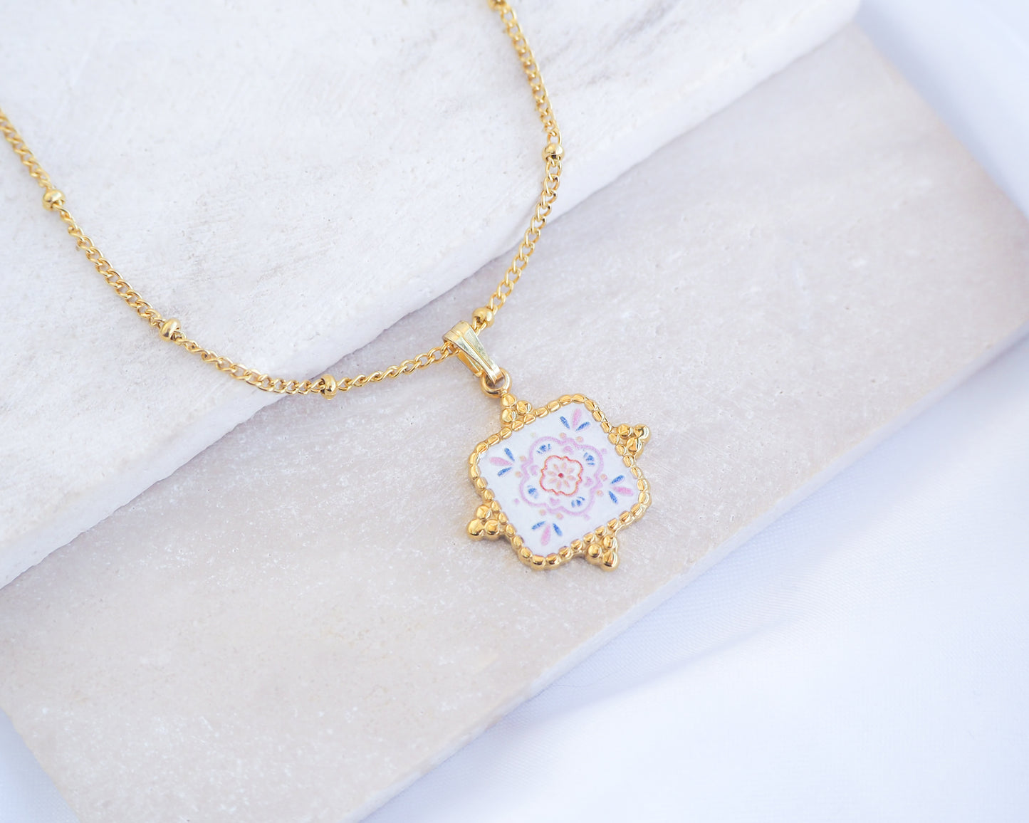 Close up of Pastel Floral Portuguese Tile Square Necklaces on stone displayed. Made with 18k gold stainless steel and white pink floral pendant inspired by the Portuguese Azulejo design pattern.