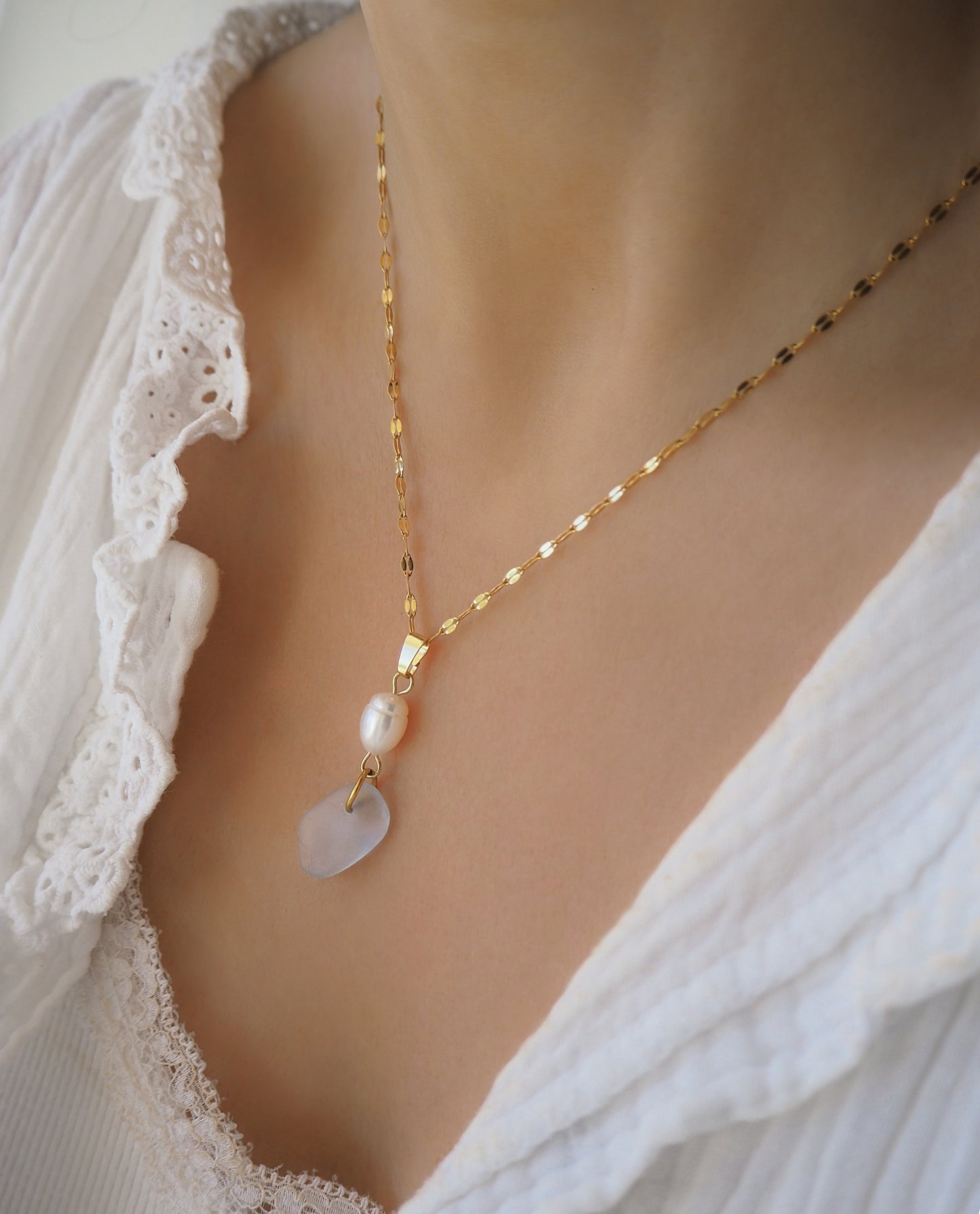 Model wearing a gold necklace with a pendant featuring a pearl and sea glass on neck with a white lace top. Necklace with a pearl and teardrop blue seaglass. Coastal jewelry with freshwater pearl necklace. Made with light blue sea glass and 18k gold stainless steel. Ocean-inspired jewelry, beach necklace, ocean treasure necklace