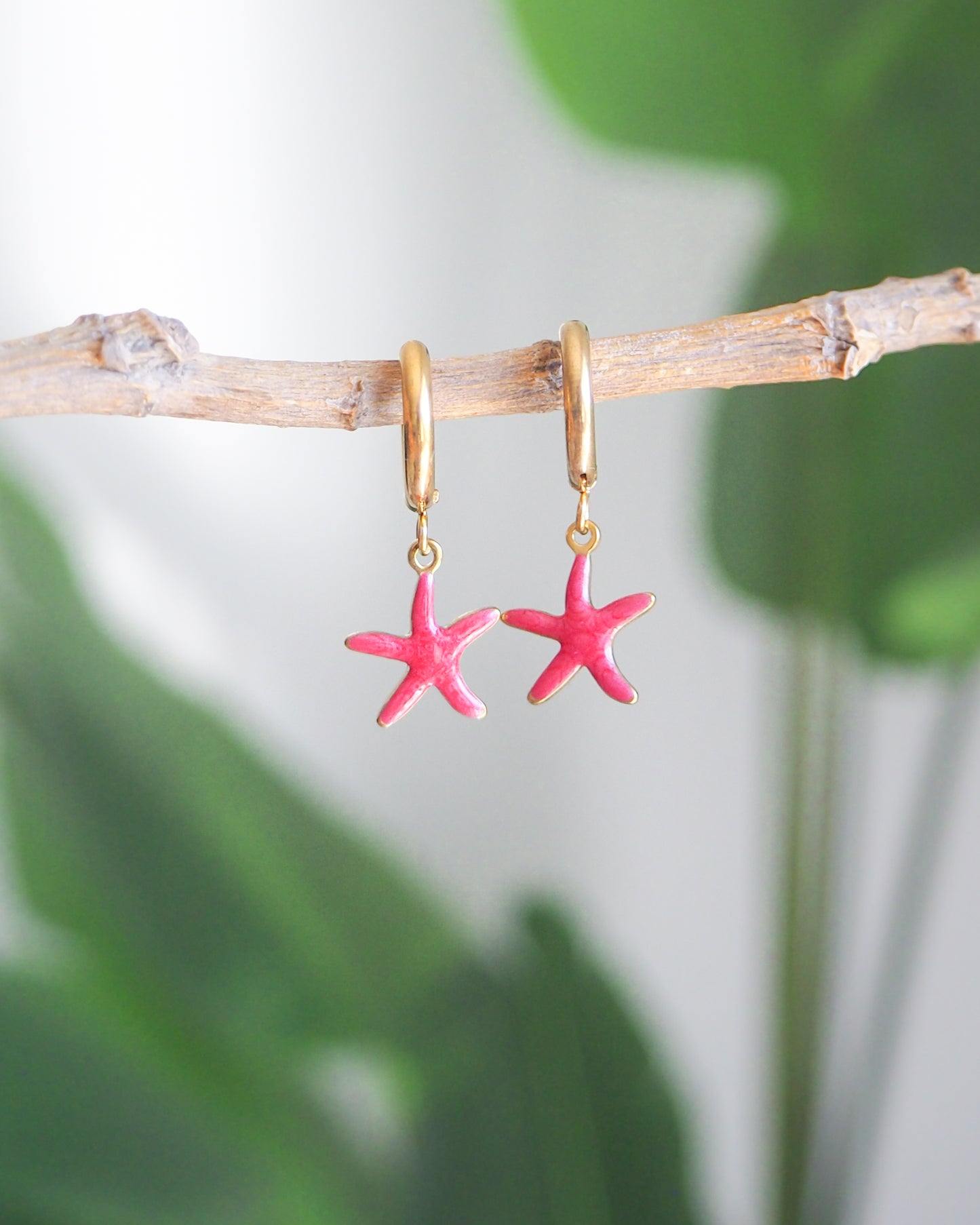 Front view of tiny starfish hoop earrings with green plant background. The starfish pendants are red. Made with 18k gold stainless steel earring hoops. Beach jewelry for summer days. Minimalistic coastal earrings. Perfect to mix and match with other earrings.