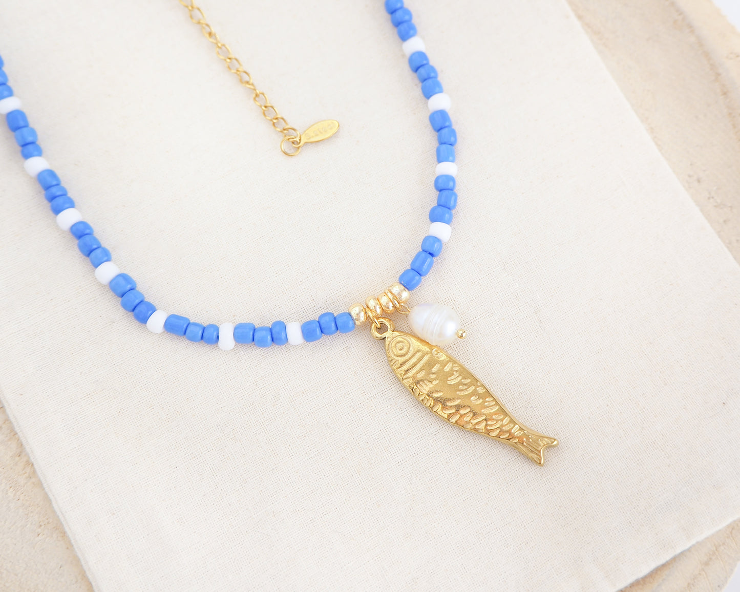 Side view of necklace with blue and white beads and a gold sardine fish pendant and freshwater pearl bead on light cotton displayed. The necklace is made with seed beads and freshwater pearls and a big gold sardine fish The sardine fish pendant is made of 18k gold stainless steel.