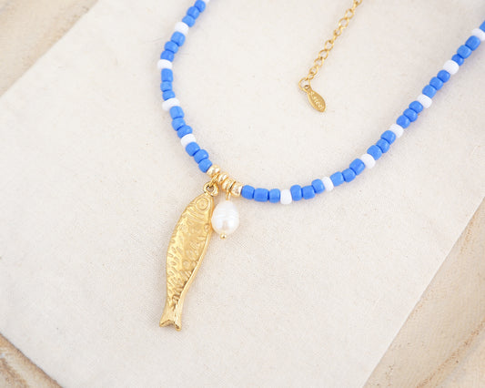 Necklace with blue and white beads and a gold fish pendant on a beige background. The necklace is made with seed beads and freshwater pearls and a big gold sardine fish The sardine fish pendant is made of 18k gold stainless steel.