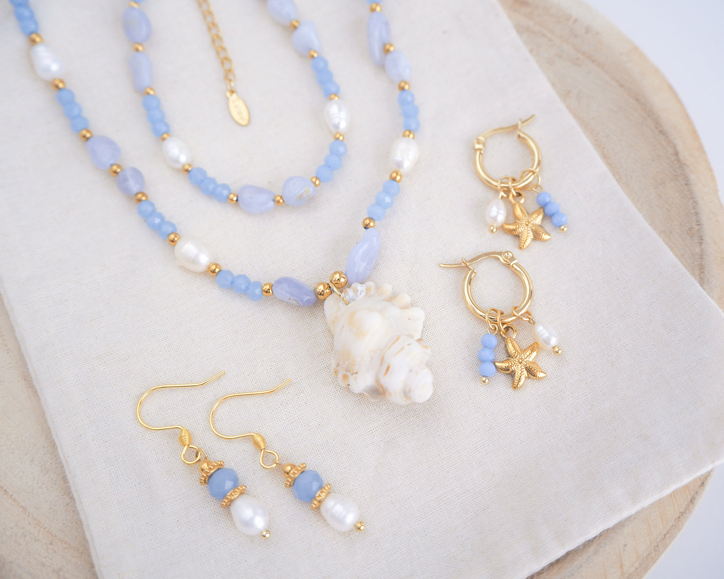 Side view of jewelry set with blue beads, gold accents, and starfish charms on a white surface. This jewelry set is part of the ocean serenity pearl collection. Handmade with a real ivory mediterranean triton shell handpicked from Portugal, blue lace agate gemstone beads, blue sparkling crystal beads, ivory freshwater pearls and 18k gold stainless steel beads.
