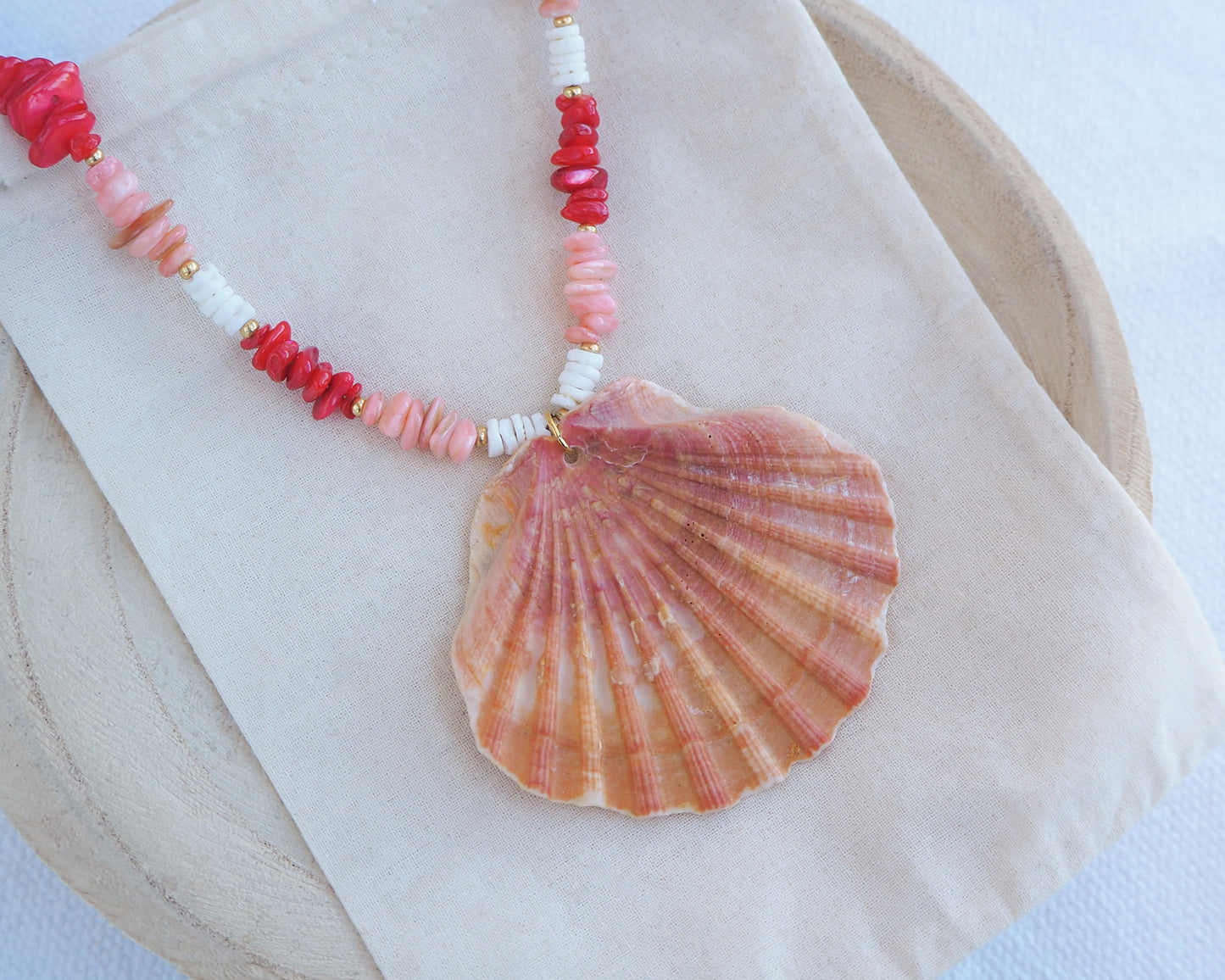 Necklace with pink and white beads and a large shell pendant on a light background. Necklace with shell pendant and red and white beads on a light blue background. Big scallop shell necklace paired with red stone beads, pink stone beads and white shell beads. The shell is a great scallop shell from Portugal and is about 7 cm. The color of the shell is orange pink and it has vertical ridges.
