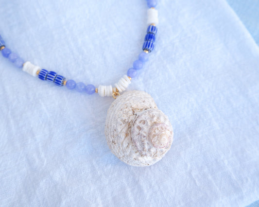 Necklace with a shell pendant and blue and white beaded chain on a light blue background. A handmade necklace with a big star shell paired with blue angelite gemstone beads, blue striped beads and white shell beads. The shell is a rough star shell from Portugal and is about 5 cm. The color of the shell is ivory white and round in shape.