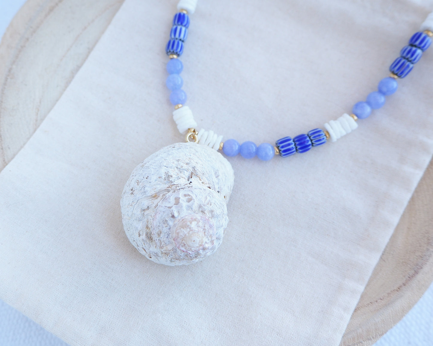 Necklace with a round white pendant and blue and white beaded chain on a light background. A handmade necklace with a big star shell paired with blue angelite gemstone beads, blue striped beads and white shell beads. The shell is a rough star shell from Portugal and is about 5 cm. The color of the shell is ivory white and round in shape.