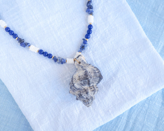 Close up of handmade necklace with a big grey murex shell paired with blue sodalite gemstone beads and white shell beads. The shell is a banded dye murex shell from Portugal and is about 8 cm. The color of the shell is grey.