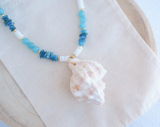 Necklace with blue and white beads and a large shell pendant on a light background. A necklace with a ivory murex shell paired with aquamarine gemstone beads and white shell beads. The shell is a banded dye murex shell from Portugal and is about 6 cm. The color of the shell is ivory bege.
