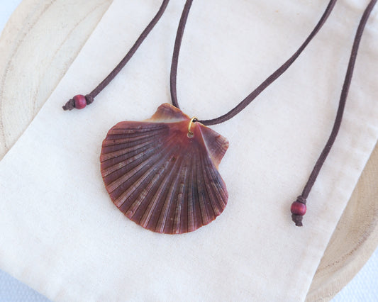 Side view of a necklace with a big scallop shell on a brown suede cord on light cotton displayed. The shell is a great scallop shell from Portugal and is about 6 cm. The color of the shell is bordeaux brown and it has vertical ridges.