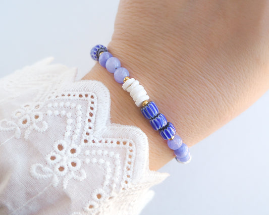 Model wearing a beaded bracelet with blue and white beads on wrist. Bracelet with blue angelite gemstones, dark blue striped beads and white shell beads displayed on a light cotton and blue background.