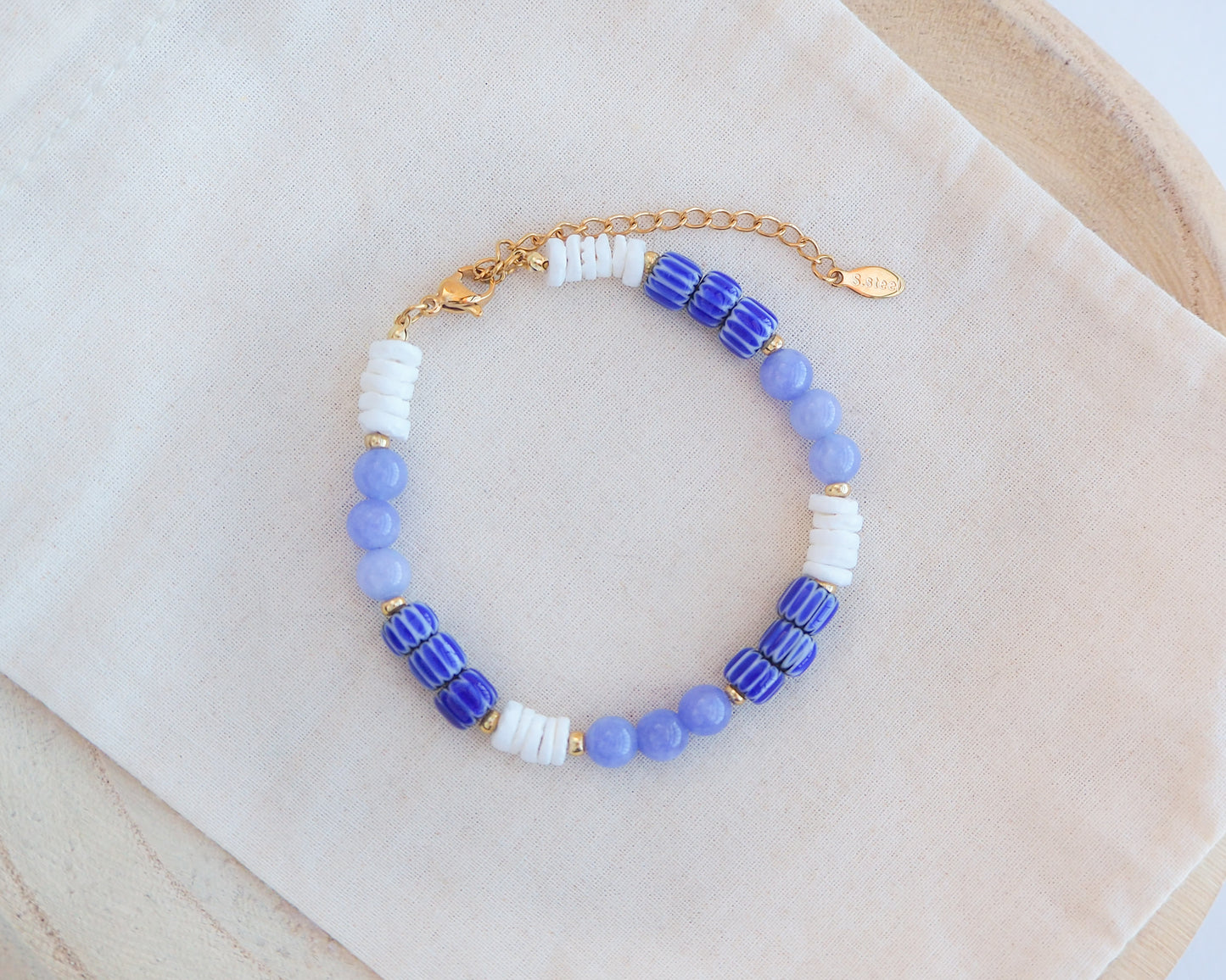 Beaded bracelet with blue, white, and gold beads on a beige fabric background. Front view of Bracelet with blue angelite gemstones, dark blue striped beads and white shell beads displayed on a light cotton .