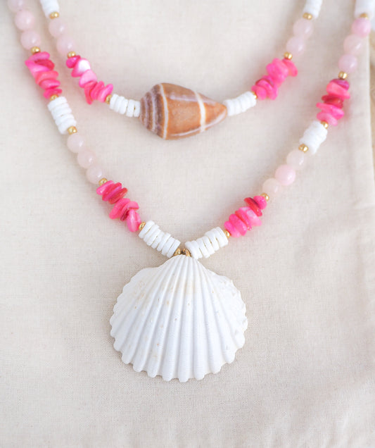 Necklace with pink beads and a white shell pendant on a light background. Necklace with pink and white beads and a shell pendant on light cotton displayed. A big white cockle shell and a necklace with a big cone shell paired with rose quartz gemstone beads, pink stones and white shell beads. The shell is a mediterranean cockle shell and Mediterranean Cone Shell from Portugal and is about 4 cm. The color of the shell is ivory and brown.