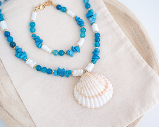 Blue and white beaded necklace with a shell pendant on a beige fabric background. A necklace with a big clam shell paired with agate gemstone beads, turquoise stones and white shell beads on cotton displayed. The shell is a mediterranean cockle shell from Portugal and is about 4 cm. The color of the shell is ivory bege. There is also a matching bracelet on the cotton dipslayed