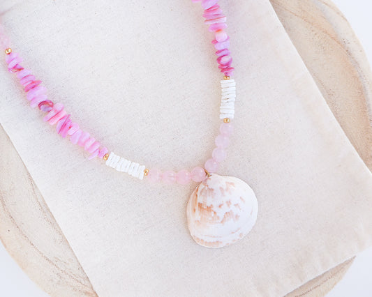 Close up of a necklace with pink and white beads and a shell pendant on light cotton displayed. A necklace with a big white cockle shell paired with rose quartz gemstone beads, soft pink stones and white shell beads. The shell is a Norwegian cockle shell handpicked from Portugal and is about 4 cm. The color of the shell is ivory white with a beige brown pattern.