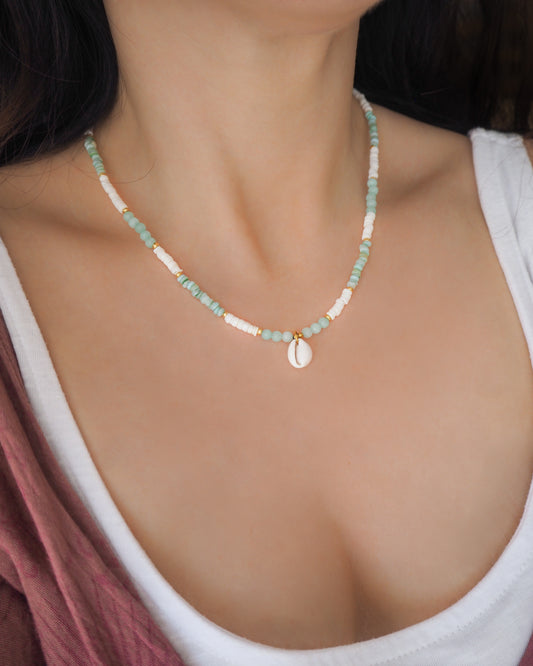 Model wearing a cowrie necklace with soft green stone beads, jade beads and white shell beads. The center piece of this necklace is a Northern Cowrie Shell, handpicked from the beach of Portugal. The shell is white and about 1 cm.