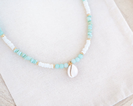 Necklace with turquoise and white beads on a light background. Close up of a cowrie necklace with soft green stone beads, jade beads and white shell beads. The center piece of this necklace is a Northern Cowrie Shell, handpicked from the beach of Portugal. The shell is white and about 1 cm.