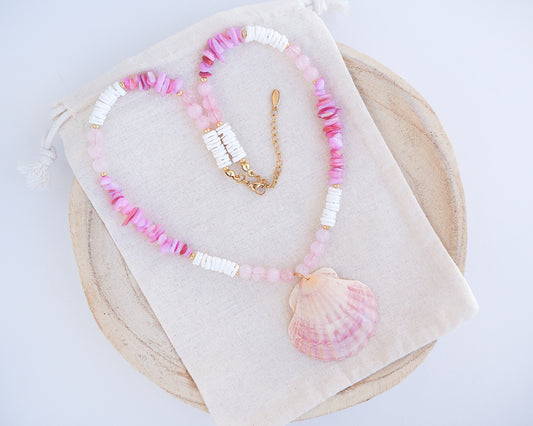 A necklace with pink and white beads and a shell pendant on light cotton displayed. A necklace with a big pink scallop shell paired with rose quartz gemstone beads, soft pink stones and white shell beads. The shell is a pilgrim's scallop shell handpicked from Portugal and is about 5 cm. The color of the shell is soft pink.