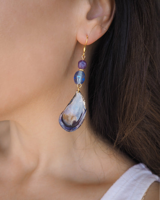 Model wearing handmade blue mussel shell earring with a gold earring hooks, purple amethyst gemstones and clear blue glass beads. The mussel shells were handpicked from the beaches of Portugal and they have a dark blue and white coloring.