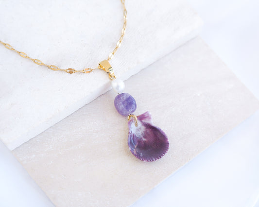 A gold chain necklace featuring a tiny scallop shell, a purple amethyst gemstone bead and a small white pearl bead displayed on a light grey stone. The shell is a variegated scallop shell handpicked from the beaches of Portugal and it has a plum bordeaux color.