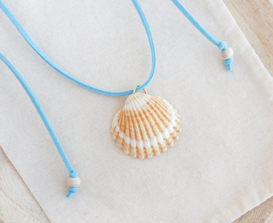 A seashell pendant necklace with a light blue cord and white beads, displayed on a textured cream fabric background. The shell is a mediterranean cockle shell from Portugal and is about 5 cm. The color of the shell is yellow white and it has vertical ridges.