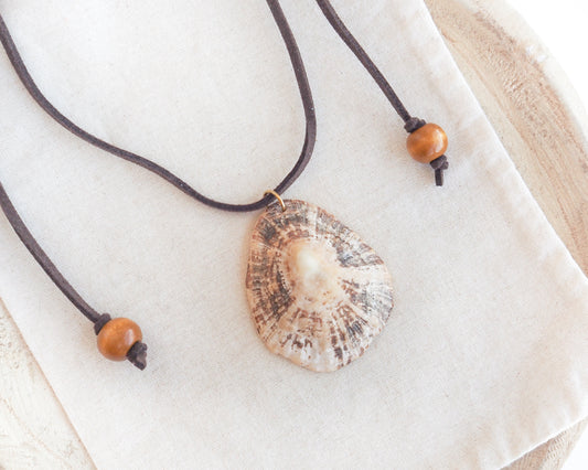 A natural limpet shell pendant necklace on a dark brown cord with wooden beads, displayed on a textured cream linen cloth. A seashell pendant necklace with a brown suede cord. The shell is a limpet shell from Portugal and is about 5 cm. The color of the shell is brown beige and it has ridges.