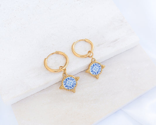 A pair of gold hoop earringd with blue and white portuguese tile pattern on stone displayed. Made with gold stainless steel and blue white center pattern that are inspired by the Portuguese Azulejo design pattern.