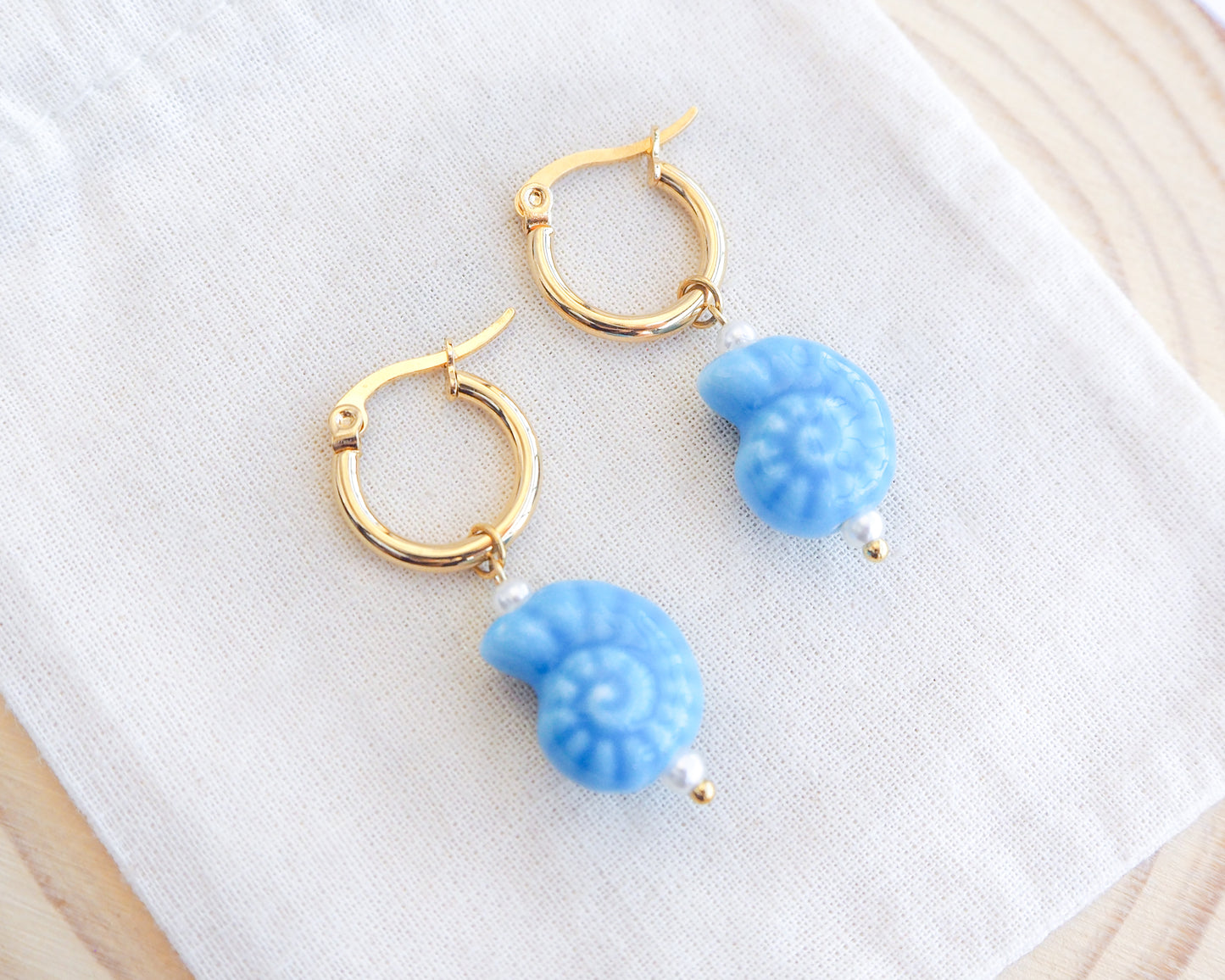 Gold ceramic seashell hoop earrings with blue nautilus seashell charms on a white cloth background