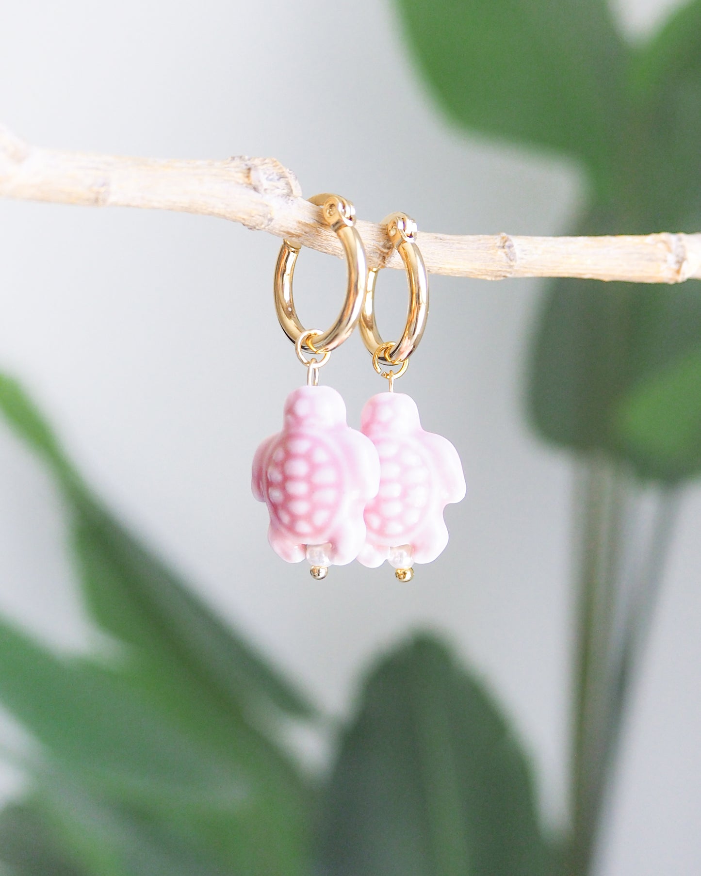 Gold hoop earrings with pink turtle charms on a branch with green leaves in the background