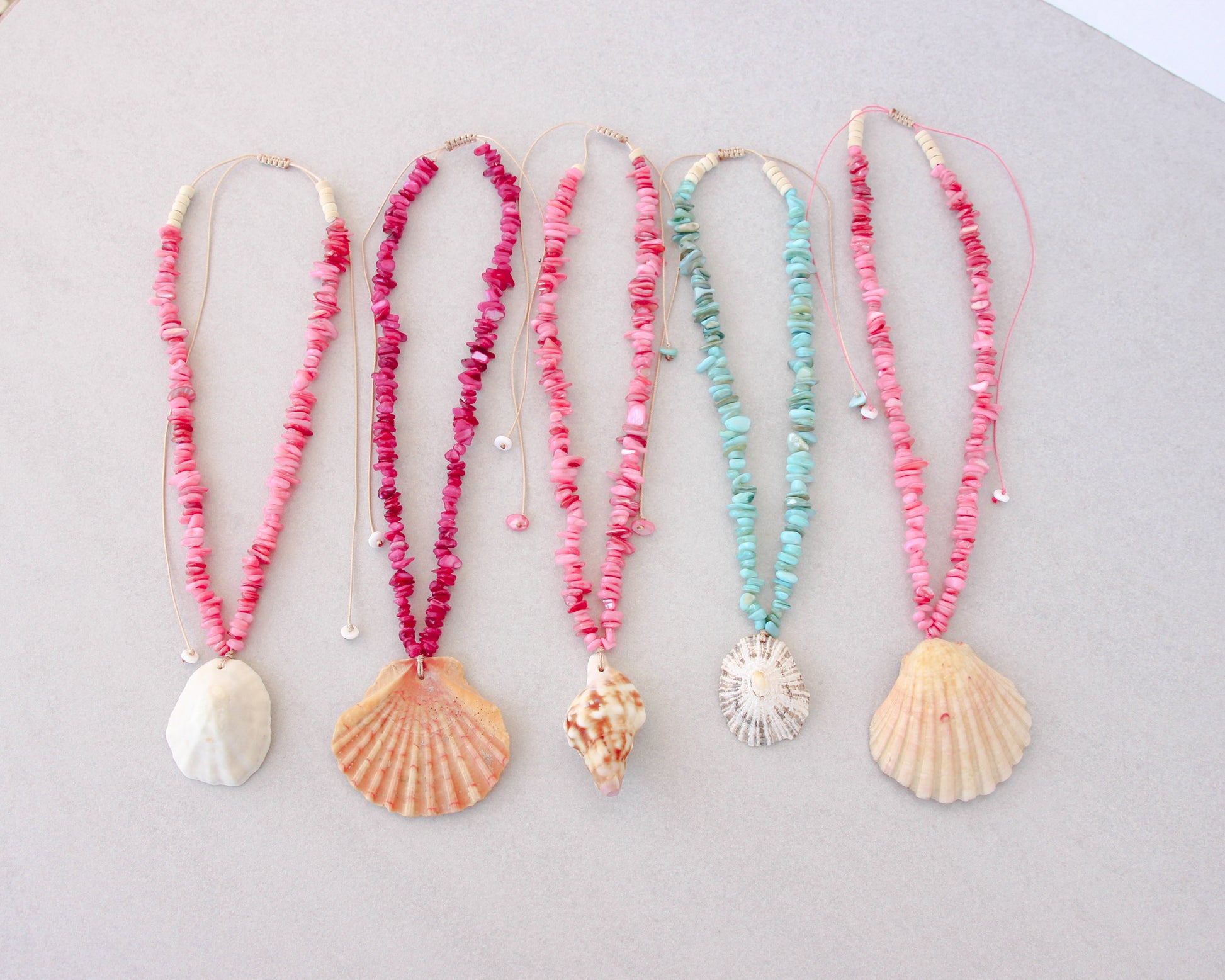 Five handmade shell bead necklaces with seashell pendants are arranged in a row on a light gray background. The necklaces feature pink, magenta, and turquoise beads with natural seashells as pendants. Portuguese shell necklaces all handpicked real big shells. 