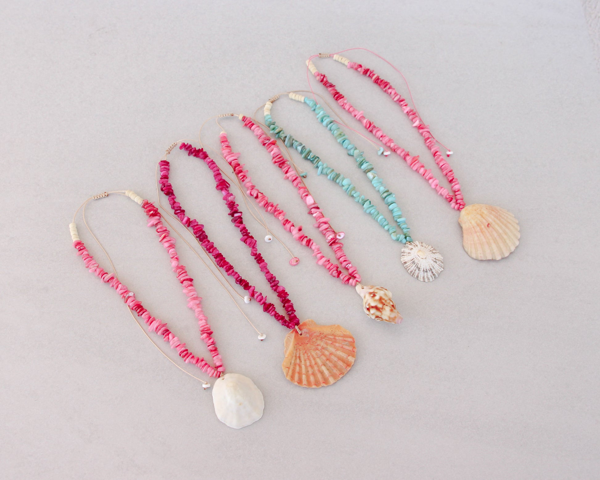 Side view of five handmade shell bead necklaces with seashell pendants are arranged in a row on a light gray background. The necklaces feature pink, magenta, and turquoise beads with natural seashells as pendants. Portuguese shell necklaces all handpicked real big shells. 
