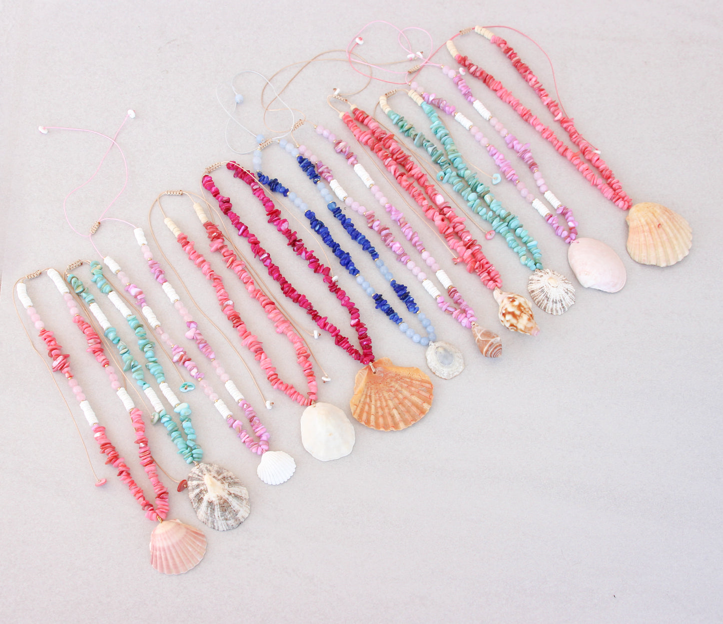 A collection of colorful handmade shell bead necklaces with seashell pendants are arranged in a row on a light gray background. The necklaces feature pink, magenta, blue and turquoise beads with natural seashells as pendants. Portuguese shell necklaces all handpicked real big shells. 