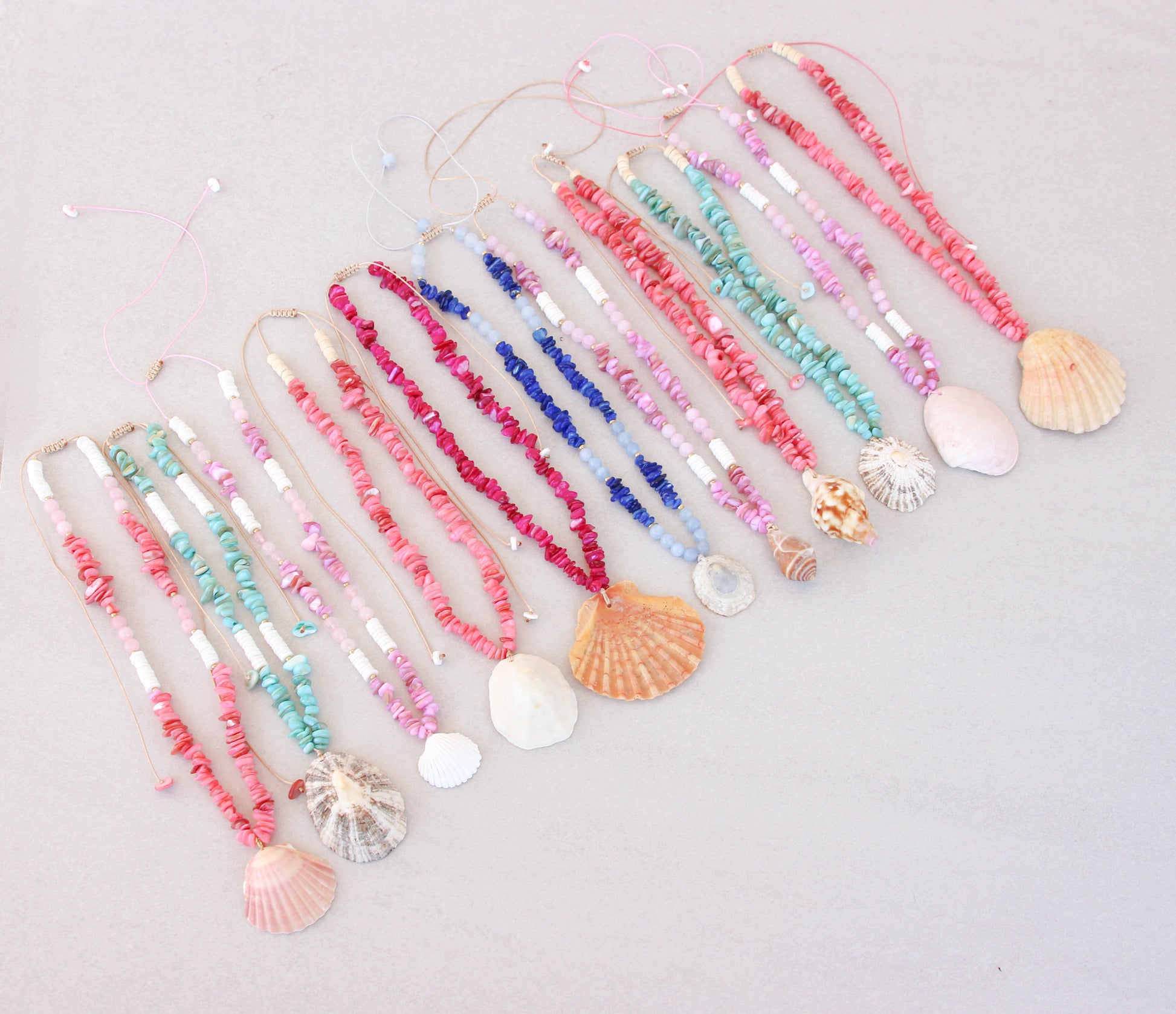 A collection of colorful handmade shell bead necklaces with seashell pendants are arranged in a row on a light gray background. The necklaces feature pink, magenta, blue and turquoise beads with natural seashells as pendants. Portuguese shell necklaces all handpicked real big shells. 