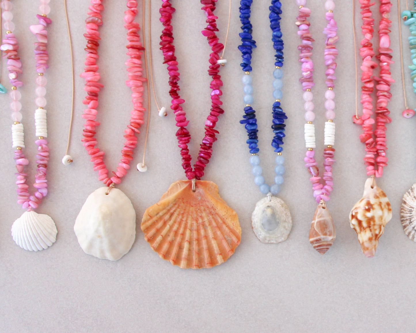 A row of colorful handmade beaded necklaces with various seashell and shell-shaped pendants, displayed against a neutral gray background.