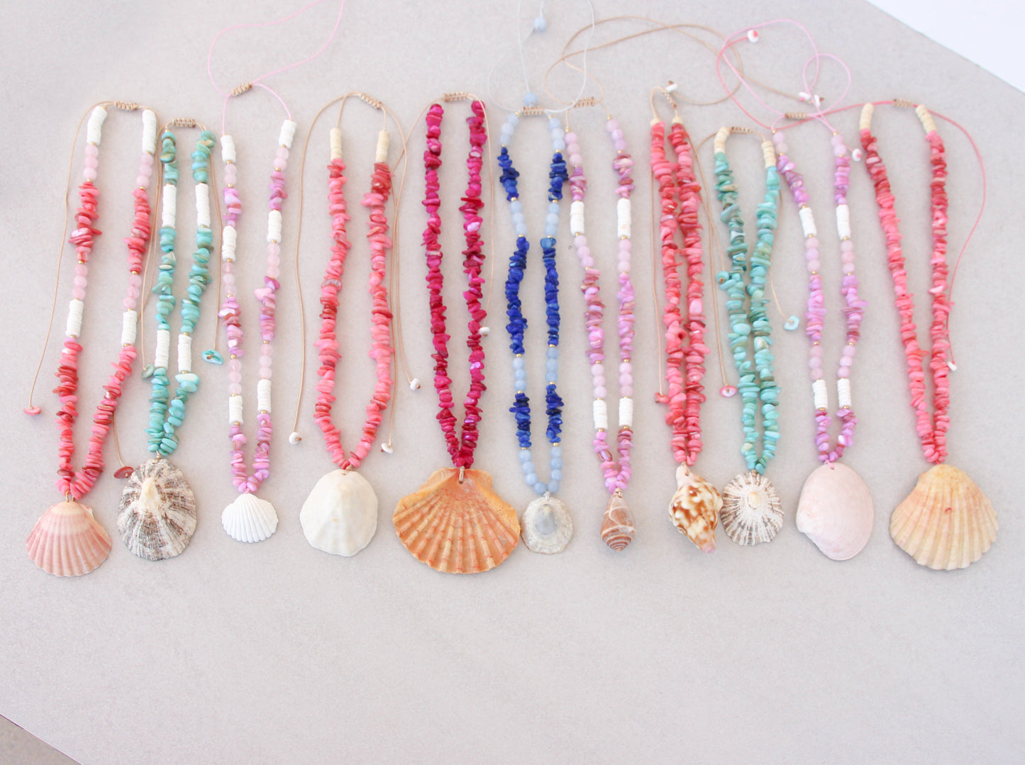A set of colorful beaded necklaces with seashell and scallop shell pendants, arranged in a row on a light beige surface. A collection of colorful handmade shell bead necklaces with seashell pendants are arranged in a row on a light gray background. The necklaces feature pink, magenta, blue and turquoise beads with natural seashells as pendants. Portuguese shell necklaces all handpicked real big shells. 