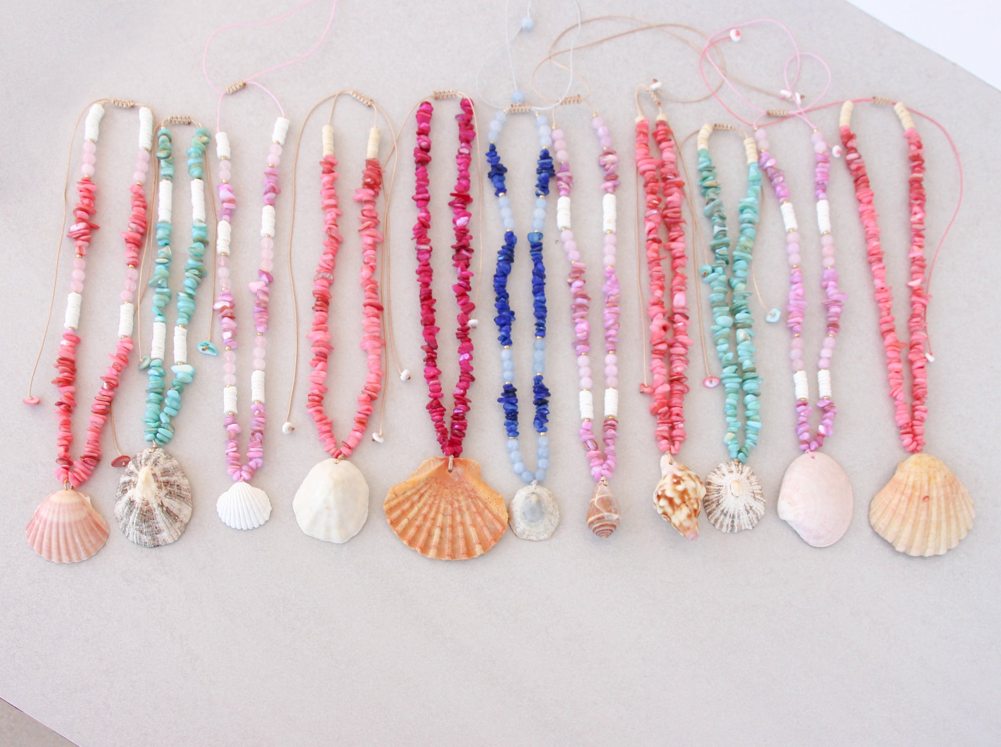A set of colorful beaded necklaces with seashell and scallop shell pendants, arranged in a row on a light beige surface. A collection of colorful handmade shell bead necklaces with seashell pendants are arranged in a row on a light gray background. The necklaces feature pink, magenta, blue and turquoise beads with natural seashells as pendants. Portuguese shell necklaces all handpicked real big shells. 