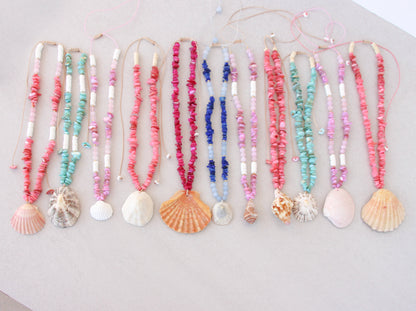 A set of colorful beaded necklaces with seashell and scallop shell pendants, arranged in a row on a light beige surface. A collection of colorful handmade shell bead necklaces with seashell pendants are arranged in a row on a light gray background. The necklaces feature pink, magenta, blue and turquoise beads with natural seashells as pendants. Portuguese shell necklaces all handpicked real big shells. 