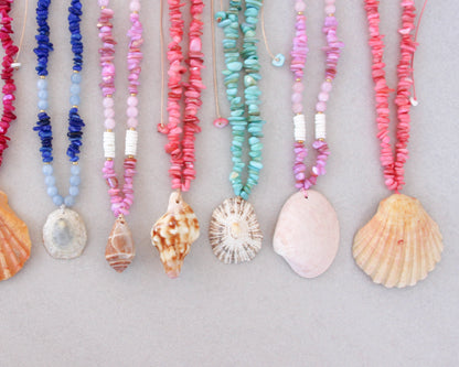 Close up of a collection of colorful handmade shell bead necklaces with seashell pendants are arranged in a row on a light gray background. The necklaces feature pink, magenta, blue and turquoise beads with natural seashells as pendants. Portuguese shell necklaces all handpicked real big shells. 