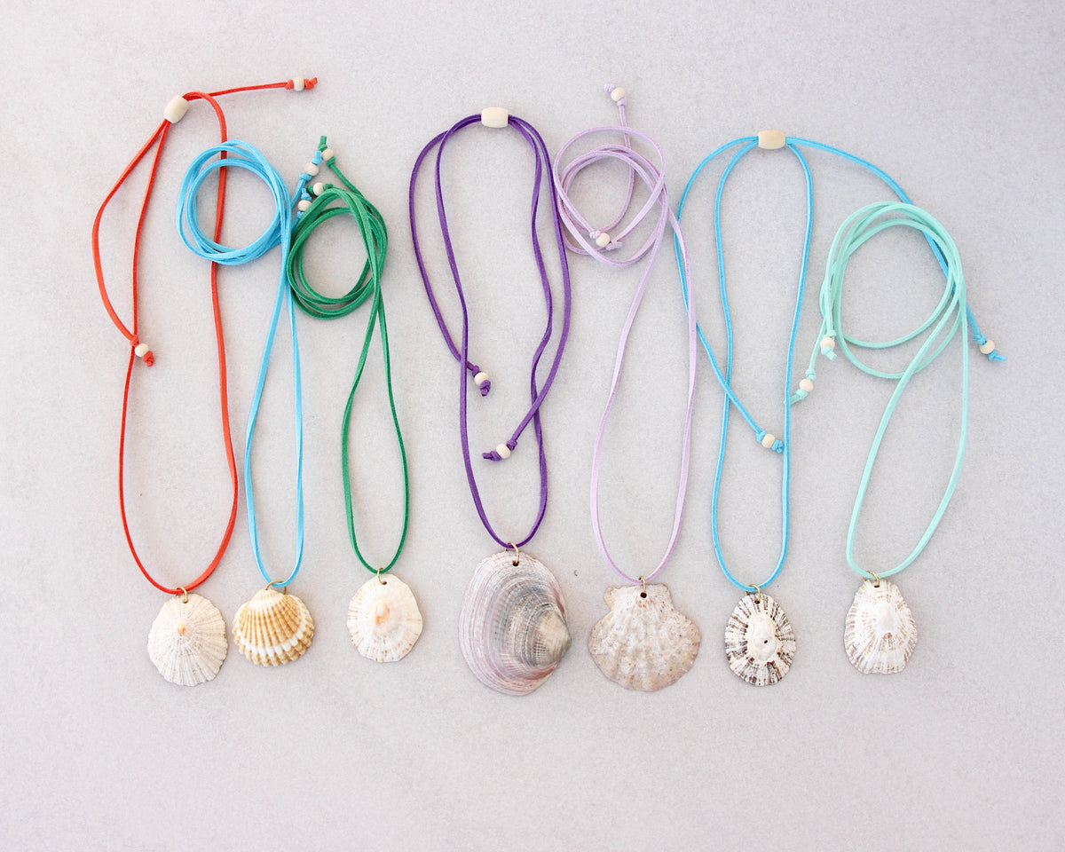 Seven seashell necklaces with colorful suede cords in red, light blue, green, purple, lavender, and mint, arranged on a light gray background.