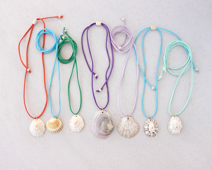 Seven seashell necklaces with colorful suede cords in red, light blue, green, purple, lavender, and mint, arranged on a light gray background.
