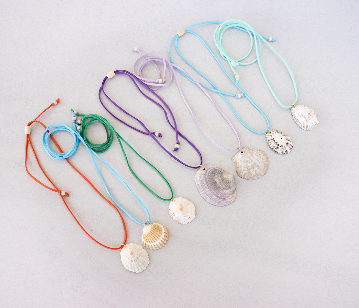 Seven adjustable suede cord necklaces with natural seashell pendants, displayed in a fan-like arrangement on a light gray background. The cords are orange, light blue, green, purple, light blue, and mint green.