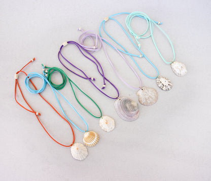 Seven adjustable suede cord necklaces with natural seashell pendants, displayed in a fan-like arrangement on a light gray background. The cords are orange, light blue, green, purple, light blue, and mint green.