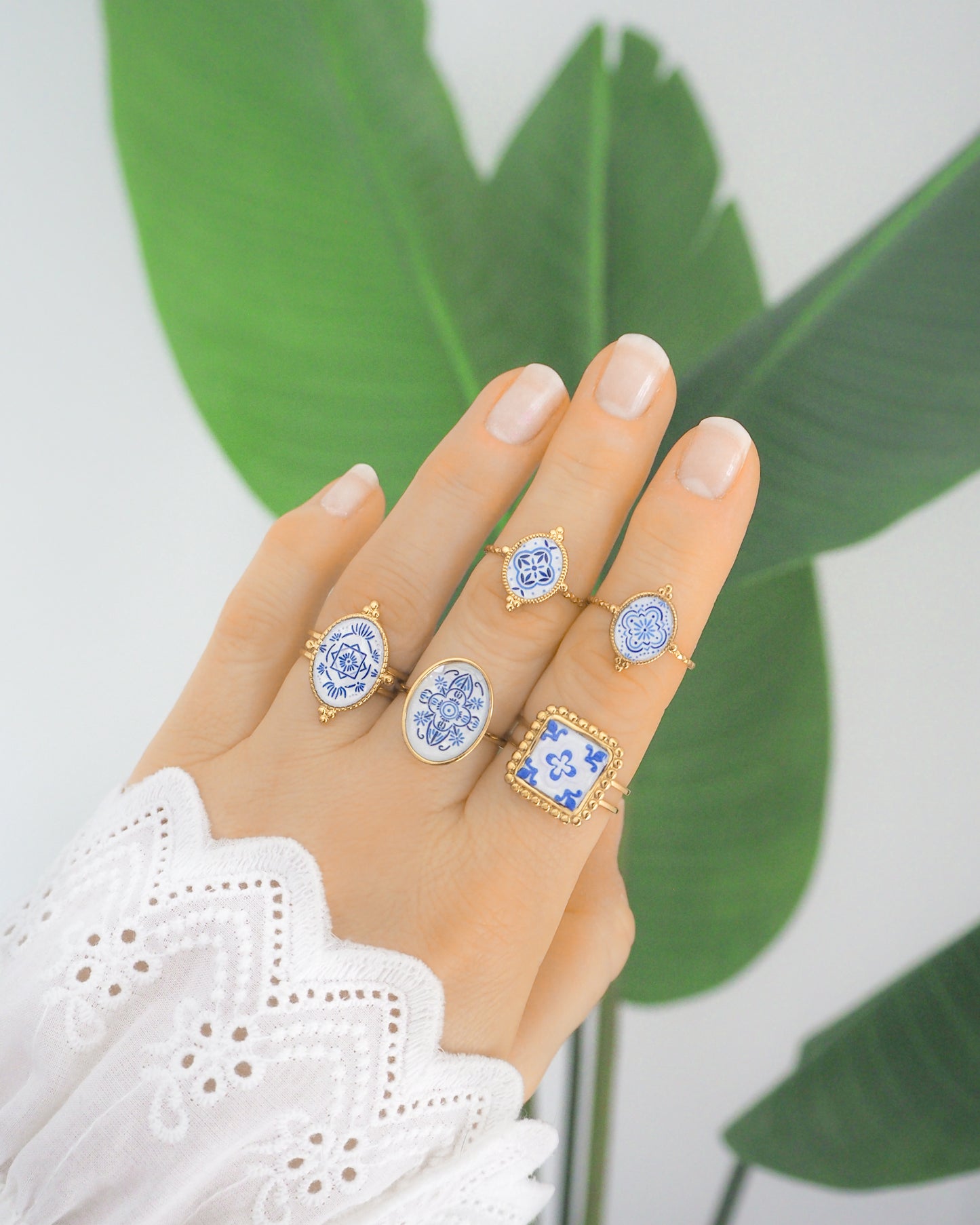 Hand wearing Blue Portuguese Tile Rings with green plant background. Rings are handmade with 18k gold stainless steel and are made with different patterns inspired by the Portuguese azulejos tiles. 