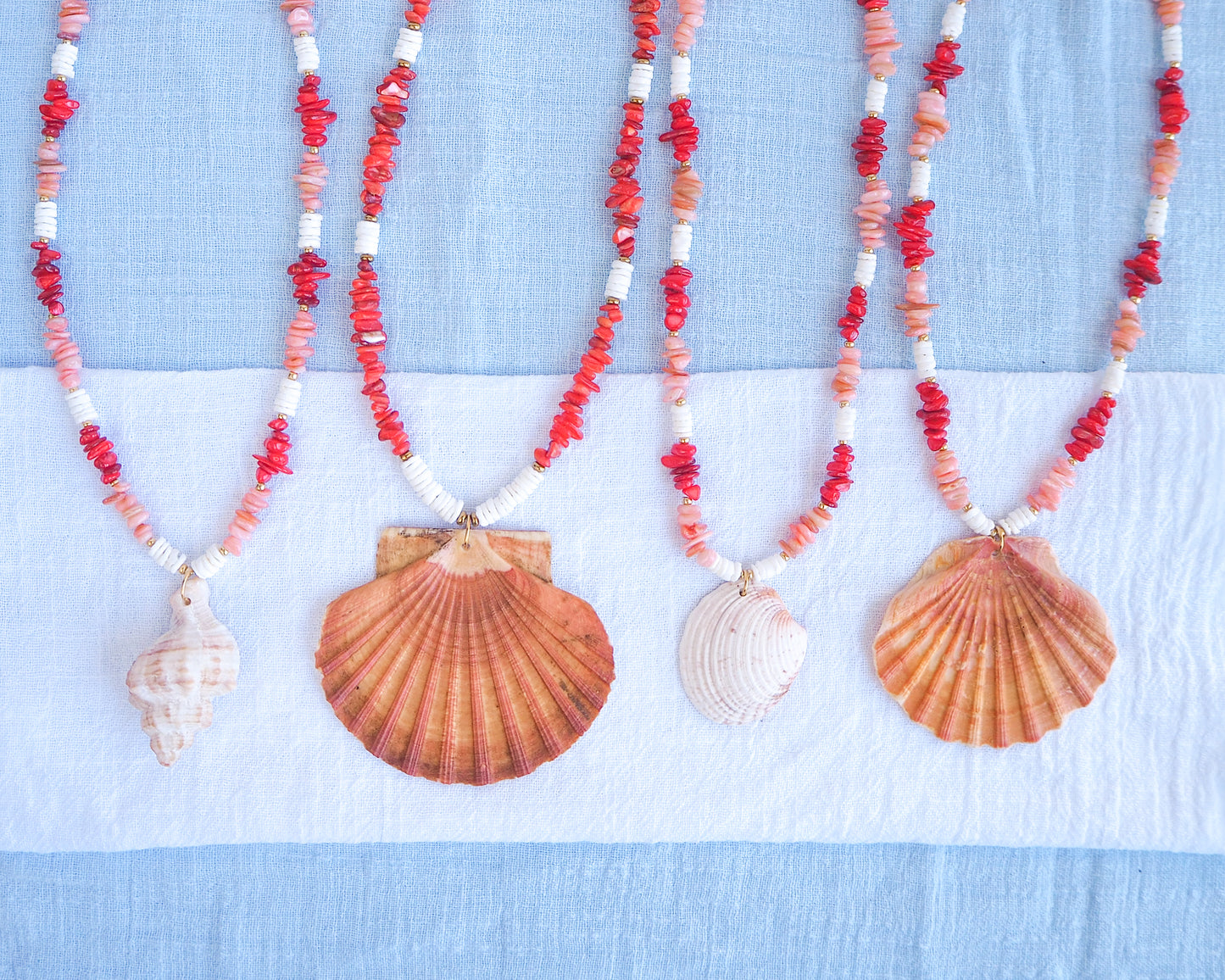 Necklaces with shell pendants and red coral stone beads on a blue and white background. A collection of big shell pendant necklaces and red and white beads on a light blue background. Big scallop shell necklace paired with red stone beads, pink stone beads and white shell beads. The shell is a great scallop shell from Portugal and is about 7 cm. The color of the shell is orange pink and it has vertical ridges. 